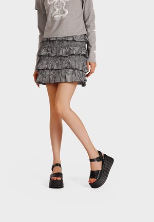 Donna che indossa una minigonna a quadri bianchi e neri a strati, una maglia grigia a maniche lunghe e sandali platform neri, in piedi con una gamba incrociata.