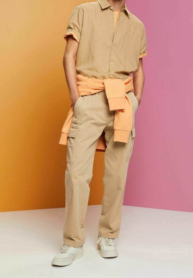 Personne portant une chemise beige à manches courtes boutonnée, un pantalon cargo beige, des baskets blanches et un sweat-shirt orange noué autour de la taille.