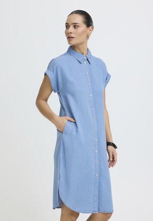 Világoskék, ujjatlan, gombos farmerruhát viselő nő, gallérral, kezét zsebében tartva, oldalra néz egy egyszerű háttér előtt.