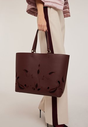 Sac fourre-tout en cuir bordeaux avec des motifs floraux découpés, deux poignées et une forme rectangulaire structurée. Texture lisse, accents en métal.