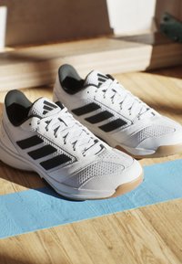 Weiße Sportschuhe mit schwarzem Drei-Streifen-Design, aus Mesh- und Synthetikmaterial, mit strukturierter Oberfläche und Gummisohle auf einem Holzboden.