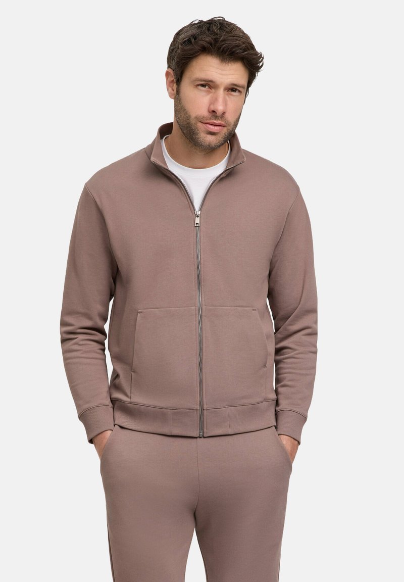 Sweatshirt zippé en tissu beige clair. Présente un col haut, des poches avant et des poignets côtelés. Texture douce avec une coupe décontractée.