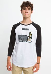 T-shirt raglan bianco e nero con un personaggio illustrato che impegna un bagaglio sovradimensionato, con il testo "Bagaglio Sovradimensionato" sopra.