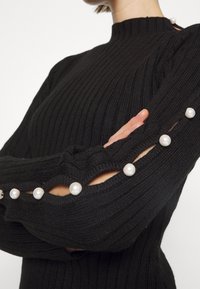 Pull noir côtelé avec manches ouvertes attachées par de gros boutons en perles blanches, porté par une personne croisant les bras.