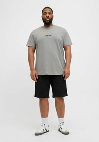 Siva pamučna majica s prednjim logotipom, u kombinaciji s crnim cargo kratkim hlačama. Model nosi bijele čarape i crno-bijele tenisice. Jednostavan, ležeran outfit.