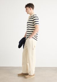 Marineblå og kremstripete polo skjorte, avslappede beige bukser, brune sko og en marineblå jakke over armen. Minimalistisk design, glatt tekstur.