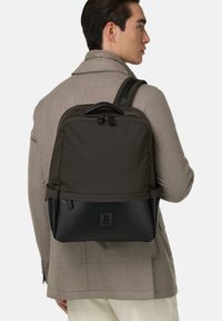 Rugzak in donkergrijze stof met een zwarte leren basis. Beschikt over dubbele ritsen en een gewatteerde handgreep, gedragen op de rug over een lichte blazer.