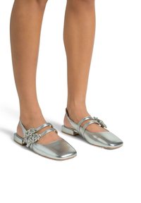 Bailarinas plateadas metálicas con puntera cuadrada, dos correas y un adorno de joya decorativa en el lateral. Presentan un tacón bajo y cuadrado.