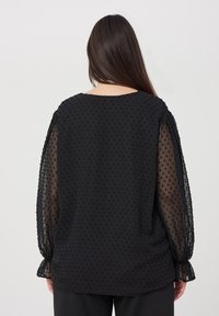 Blusa nera con maniche trasparenti a pois, scollatura rotonda e vestibilità morbida. Presenta polsini arricciati e una texture liscia.