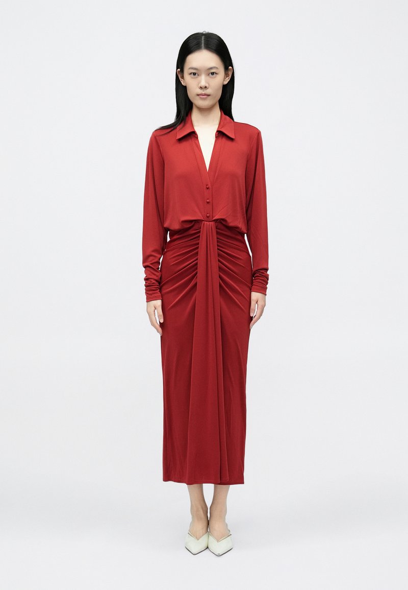 Piros jersey ruha gombos gallérral, hosszú ujjakkal és húzott elülső résszel. Laza fazonú, közép-vádli hosszúságú.