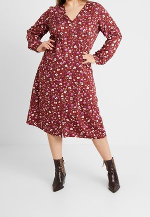 Vrouw draagt een rode midilengte jurk met lange mouwen en knopen, met een wit en paars bloemenprint, gecombineerd met donkerbruine enkellaarzen.