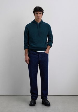 Giovane uomo in piedi contro un muro semplice, indossa un hoodie blu scuro, jeans blu scuro e scarpe nere con una mano in tasca.