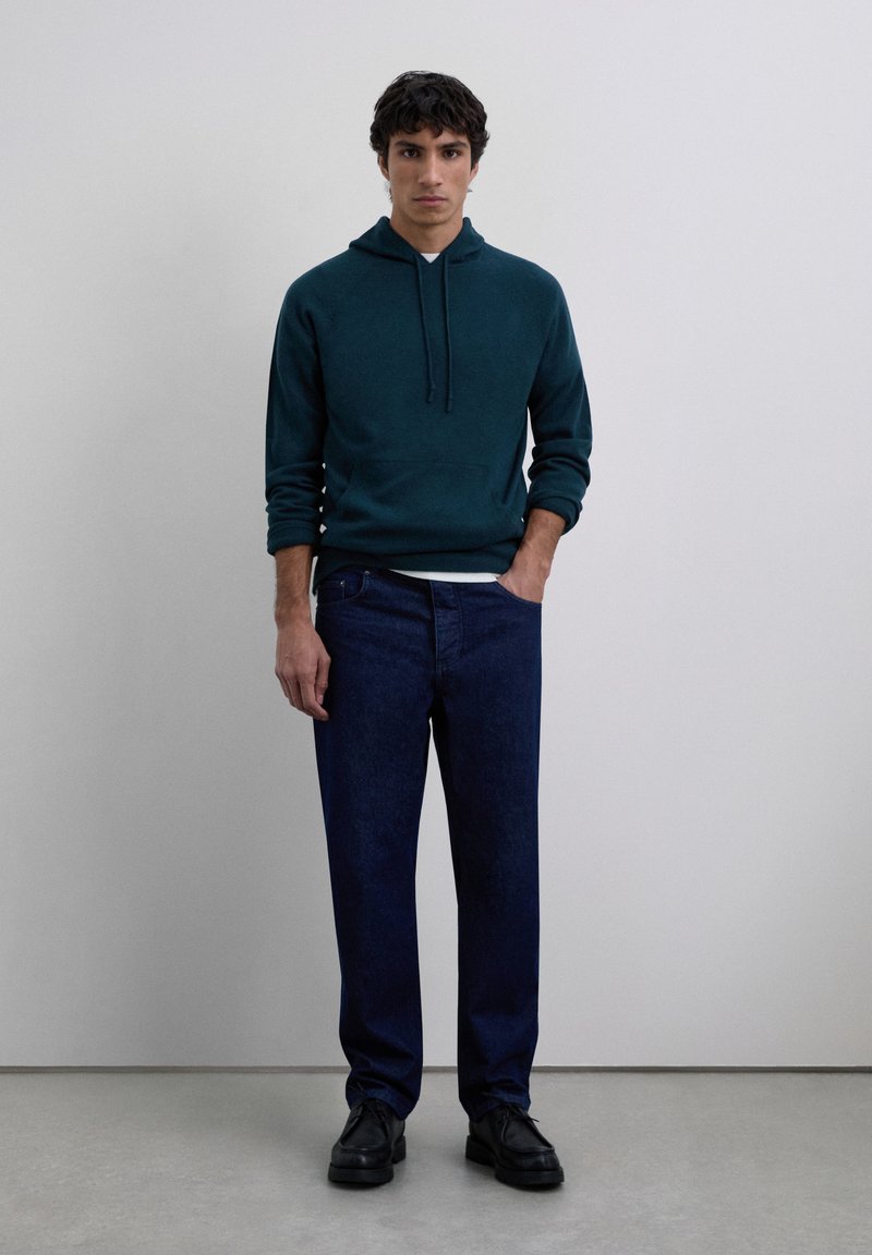 Ung man som står mot en enkel vägg, iklädd en mörkt teal hoodie, mörkblå jeans och svarta skor med ena handen i fickan.