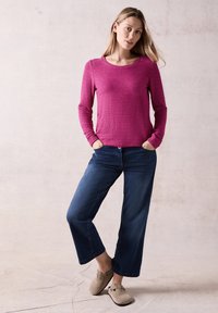 Top a maniche lunghe fucsia con righe testurizzate, abbinato a jeans in denim blu scuro a gamba larga e scarpe slip-on beige. Sfondo semplice.