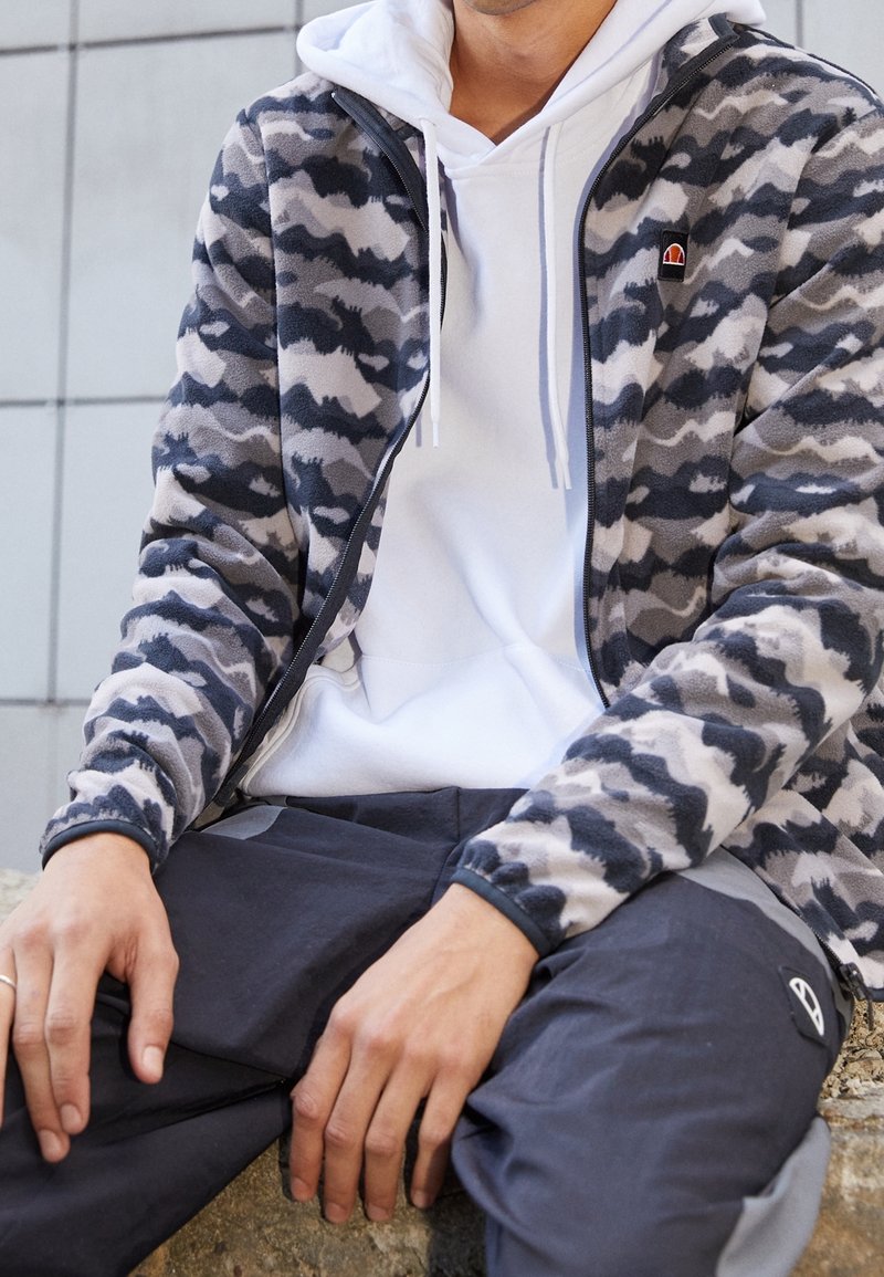 Personne portant un sweat à capuche blanc sous une veste zippée camouflage grise et noire et un pantalon foncé, assise avec les mains posées sur les genoux.