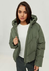 Olive green puffer jacket with a hood, featuring a zip closure and ribbed cuffs, worn over a white T-shirt and gray pants.