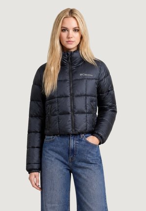 Giovane donna con giacca imbottita nera Columbia e jeans blu, in piedi con una mano in tasca davanti a uno sfondo semplice.