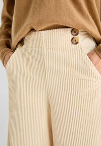 Beige fløjlsbukser med lodret ribbet tekstur, der har to brune knapper i taljen og sidelommer. En hyggelig striktrøje er delvist synlig.