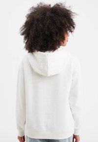 Sudadera blanca con cordón, textura suave y puños de canalé. Presenta un bolsillo canguro y un corte relajado, combinada con jeans claros.