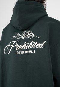 Persona che indossa una felpa con cappuccio verde scuro con testo ricamato bianco "Proibito 10119 Berlino" e un piccolo ramo sopra il testo sulla schiena.