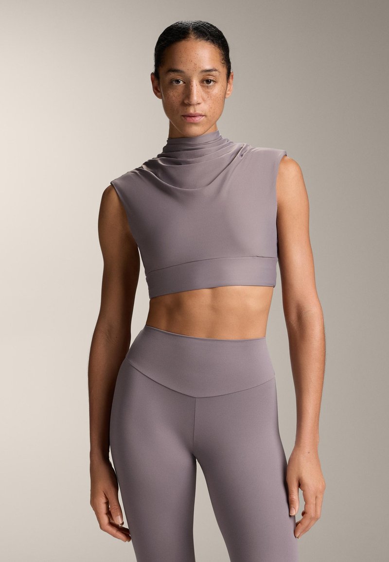 Donna con i capelli raccolti e lentiggini, che indossa un top crop senza maniche di lavanda con collo alto e leggings a vita alta coordinati, su uno sfondo neutro.