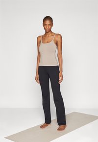 Beige tílko s tenkými ramínky a kulatým výstřihem, doplněné černými zvonovými kalhotami. Modelka stojí na světle zbarvené jóga podložce.