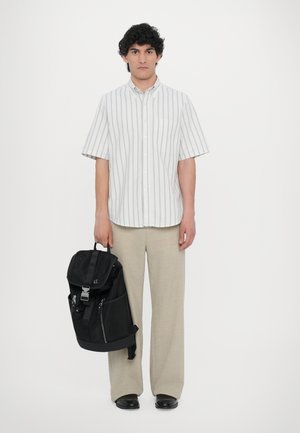 Zaino nero con più tasche e chiusura a fibbia, indossato da una persona in camicia a righe con bottoni e pantaloni larghi di colore chiaro.