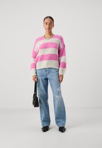 Maglione chunky a maglia con strisce rosa e bianche, abbinato a jeans chiari strappati. Una borsa nera e scarpe nere a punta completano il look.