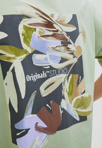 Light green t-shirt featuring a bold floral graphic in shades of brown, blue, and cream with "Originals™ STUDIO" text in the center.