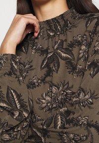 Brun blommig blus med en rynkad smockad halsringning och manschetter. Har intrikata svarta blommönster på ett mjukt, texturerat tyg.