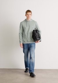 Een jonge man in een lichtgroene hoodie en blauwe spijkerbroek loopt terwijl hij een zwarte sporttas in zijn linkerarm houdt.