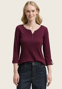 Frau mit hellblonden Haaren, die ein burgunderfarbenes langärmliges Shirt mit Knöpfen und dunkelblaue Jeans trägt, steht lächelnd vor einem schlichten Hintergrund.