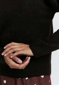 Un pull en tricot noir avec une finition texturée et des poignets côtelés, porté sur une jupe bordeaux à pois blancs, les mains montrées dans une position détendue.
