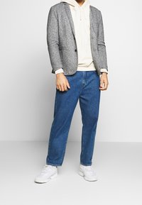 Grauer Blazer aus Wollmischung über einem beigen Hoodie, kombiniert mit locker sitzenden blauen Denim-Jeans und weißen Sneakern, zeigt einen Lagen-Look.