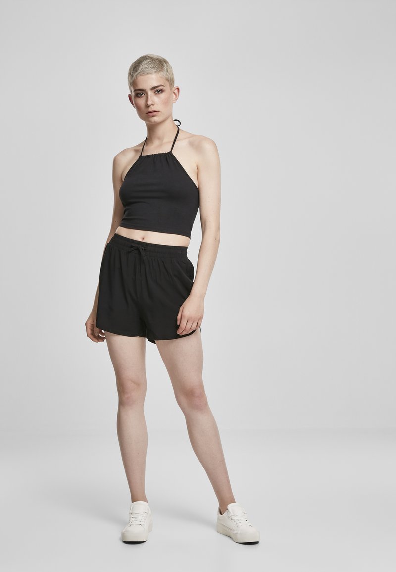 Crni halter kropped top u kombinaciji s crnim šortsevima s vezicom, s ležernim krojem. Model nosi bijele tenisice, pozira pred svijetlom pozadinom.