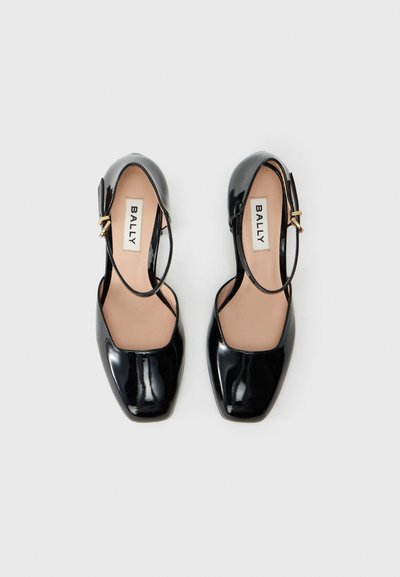 Sapatos Mary Jane pretos brilhantes com bicos quadrados e fivelas douradas, rotulados "BALLY" por dentro, vistos de cima sobre um fundo branco.