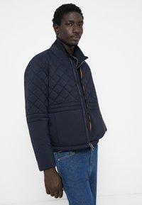 Veste matelassée bleu marine avec un col montant, un accent de fermeture éclair orange et des détails de piping horizontaux. Portée avec un jean bleu.