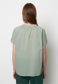 Donna con lunghi capelli castani, che indossa una blusa verde chiaro a maniche corte e pantaloni verde scuro, in piedi contro uno sfondo bianco uniforme, voltata di spalle.