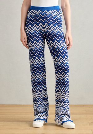 Persoon die blauwe en witte zigzaggende wijde broek draagt met een blauwe tailleband en witte sneakers op een houten vloer.