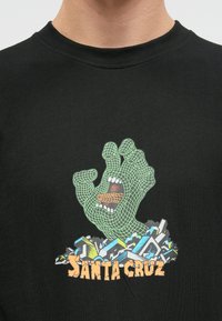Melnas kokvilnas T-krekls ar zaļu stiegru roku atvērtām mutēm, novietots pārkrāsotā grafiskā fona un ar tekstu "SANTA CRUZ".