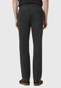 Osoba oblečená v černých rovně střižených kalhotách a černých botách s hnědými podrážkami, stojící proti jednobarevnému světle šedému pozadí.