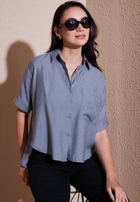 Camicia azzurro chiaro a maniche corte, con taschino sul petto, vestibilità leggermente ampia, realizzata in un tessuto liscio, abbinata a pantaloni neri.