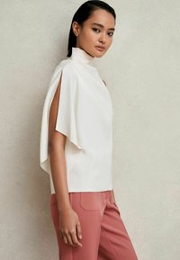 Top alb cu guler înalt și mâneci ample, purtat cu pantaloni roz skinny. Material neted, design minimalist și linii elegante.