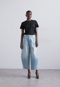 T-shirt preto de manga curta com logótipo, combinado com jeans de ganga oversized de cor azul claro. Modelo a usar sapatos de salto altos pretos, em pé contra um fundo neutro.