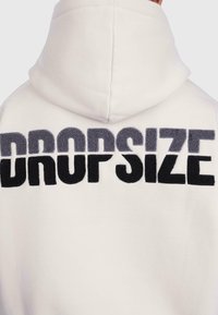 Back view of white hoodie with raised textured letters spelling "Drop Size" across the upper back under the hood.