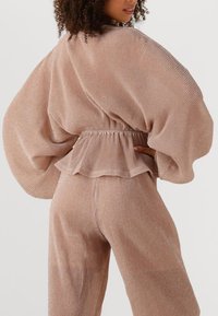 Geribbelde, metallic rosegouden tweedelige outfit met brede mouwen, elastische taille en gelaagde ruffeldetails. Zachte, gestructureerde stof.