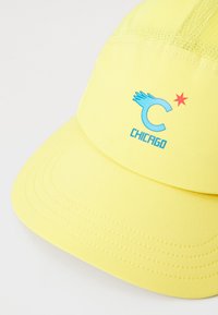 Gelbe Kappe aus Stoff, mit einem blauen "C"-Logo mit Flügeldetail und einem roten Stern, strukturiertes Design und genähte Akzente.