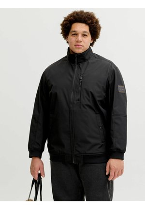 Homme aux cheveux bouclés portant une veste noire zippée et un pantalon sombre, tenant des poignées de sac noires sur un fond clair uni.