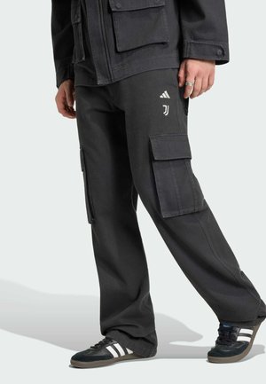Osoba ubrana w luźne czarne spodnie typu cargo z logo Adidas i Juventusu, czarną kurtkę oraz czarne buty sportowe Adidas z białymi paskami.