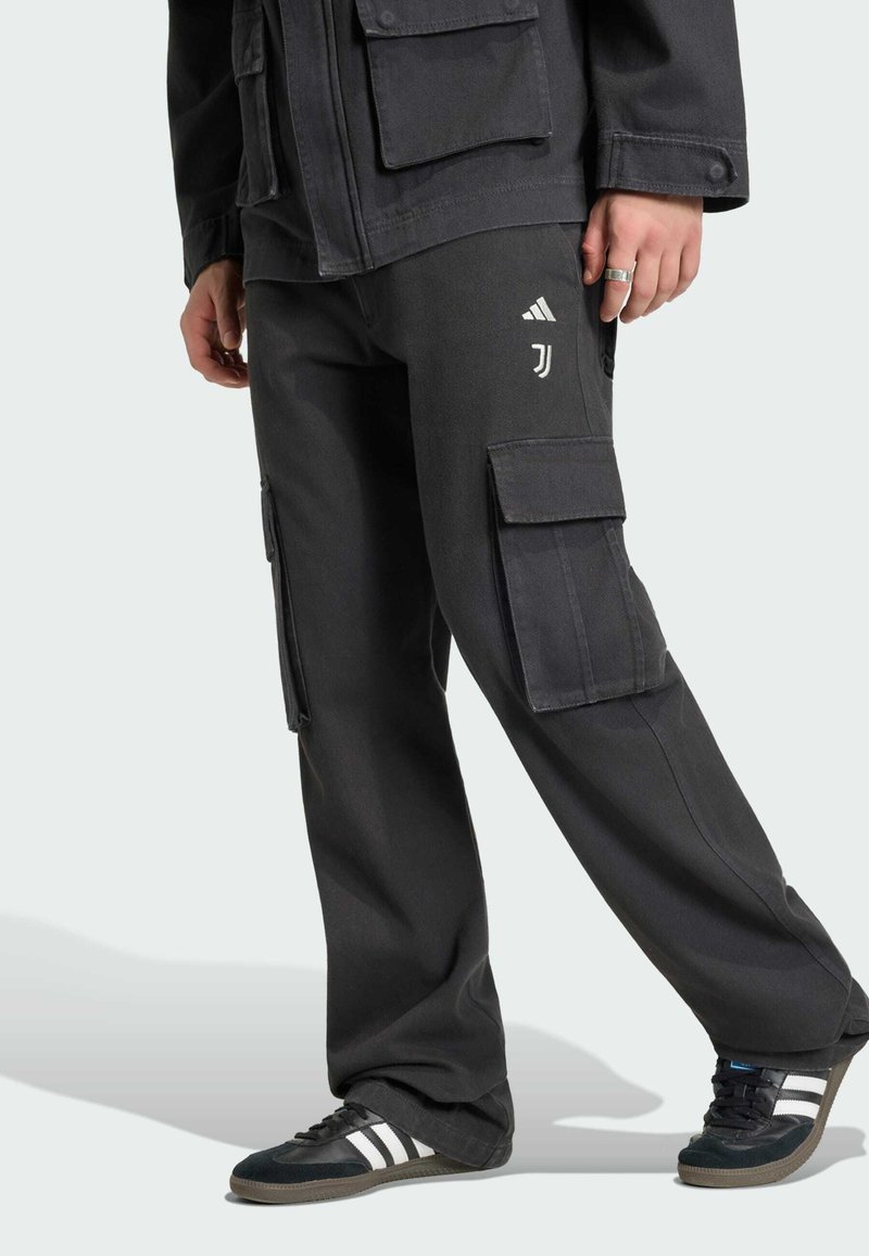 Personne portant un pantalon cargo noir ample avec les logos Adidas et Juventus, une veste noire et des baskets noires Adidas avec des bandes blanches.